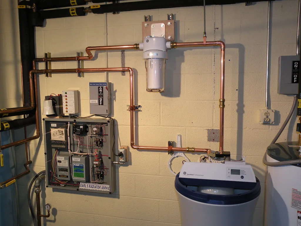 Basement with copper plumbing along a block wall, a cartridge filter housing mounted on the wall and a cabinet-style water softener with a digital display standing on the floor.