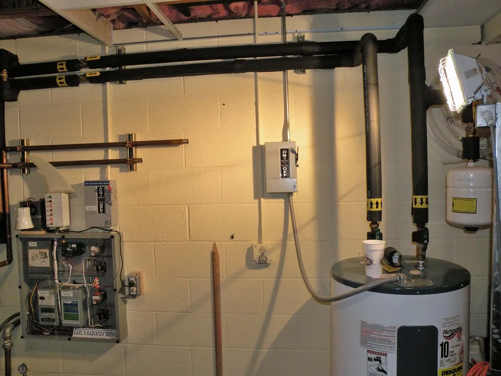 Unfinished basement with a water heater, an electrical panel and exposed pipes along a block wall, with open floor space in front.