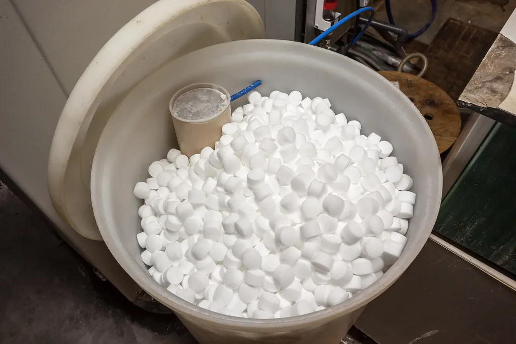 Top-down view into a round brine tank filled with white softener salt pellets, with a vertical brinewell tube at the back.