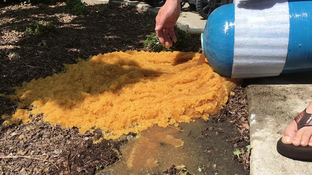 A blue resin tank tipped on its side outdoors, with a large pile of amber resin beads poured out onto the ground.